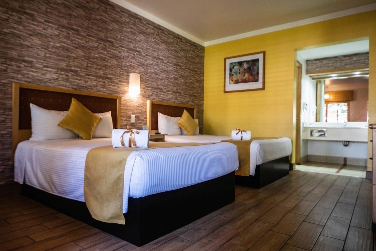 A hotel room with two beds, a brick wall, a yellow accent wall, a painting, and a bathroom with a mirror and sink.