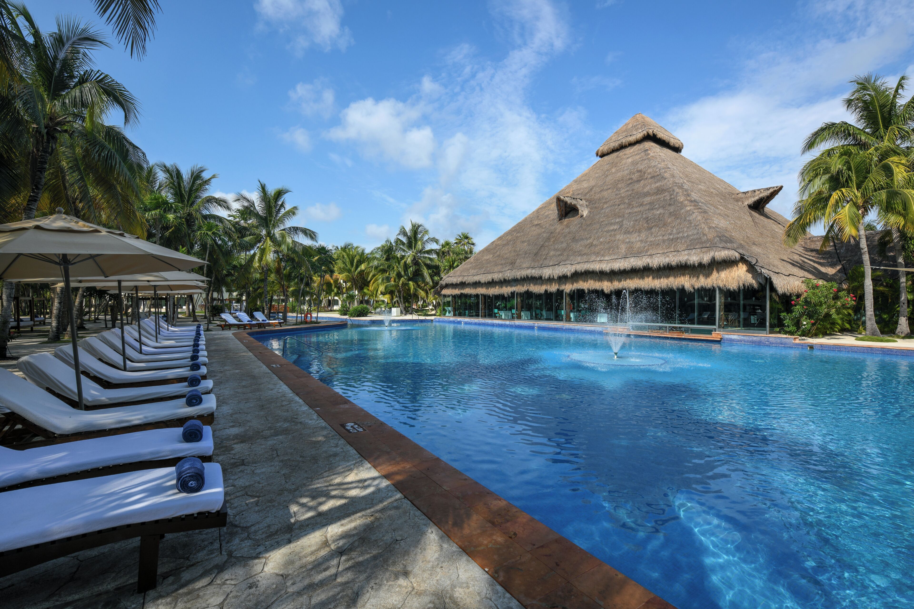 A swimming pool with lounge chairs, a thatched-roof structure, and palm trees.