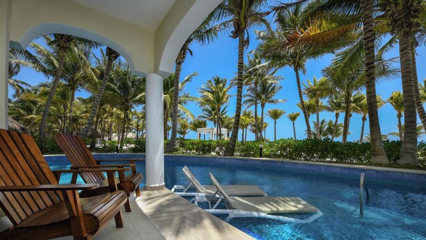 A pool area with wooden lounge chairs and a clear view of palm trees and a building in the background. - El Dorado Royale, All & More Inclusive Adults Only (Playa del Carmen)