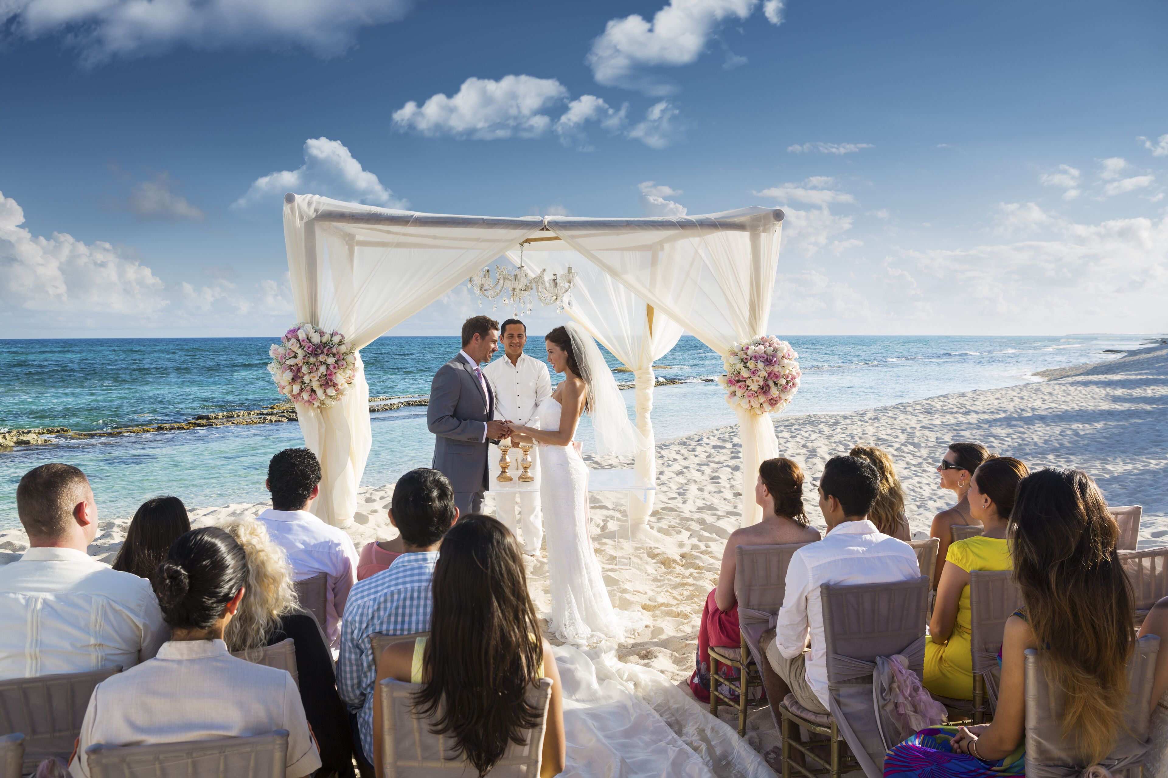 Una ceremonia de boda en la playa, con un arco blanco y los invitados sentados en sillas mirando a la pareja.