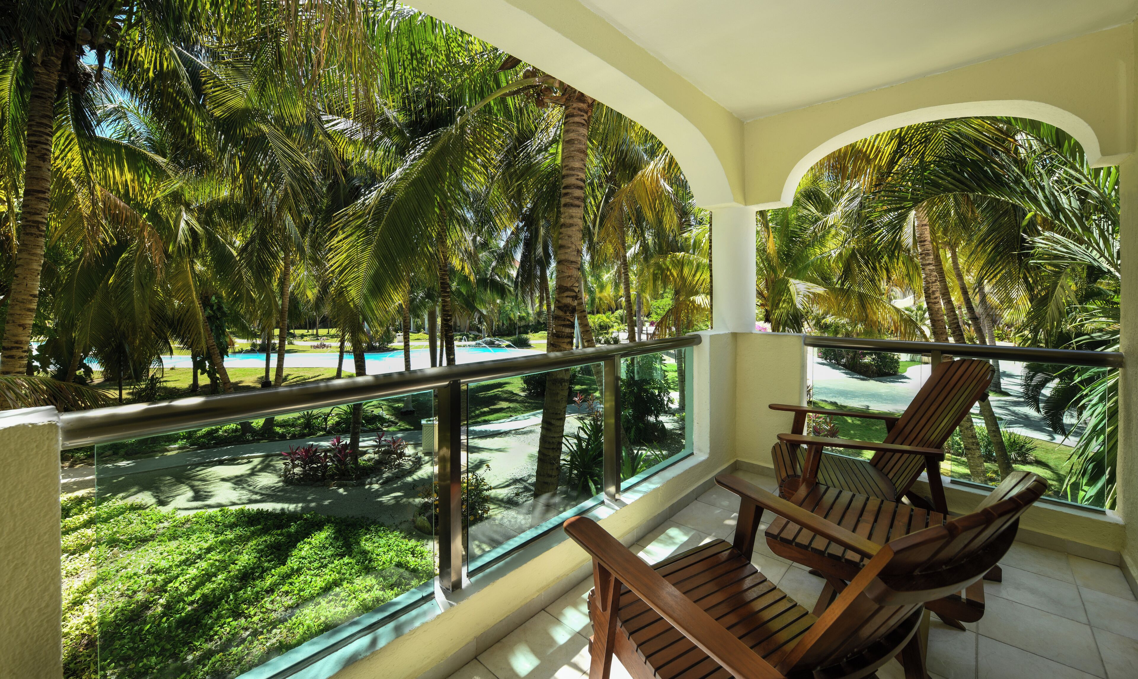 A balcony with wooden chairs, a glass railing, and a view of a swimming pool surrounded by palm trees and greenery.
