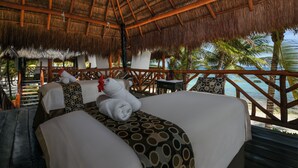A thatched-roof room with two beds, a wooden deck, and a view of the ocean and palm trees.