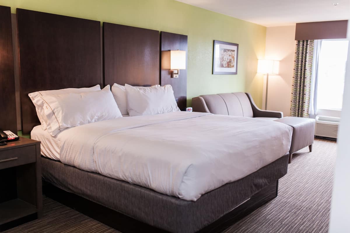 Una habitación de hotel con una cama grande, una mesita de noche con teléfono, una silla, una lámpara y una ventana con cortinas.