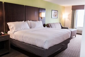 A hotel room with a large bed, a bedside table with a phone, a chair, a lamp, and a window with curtains.