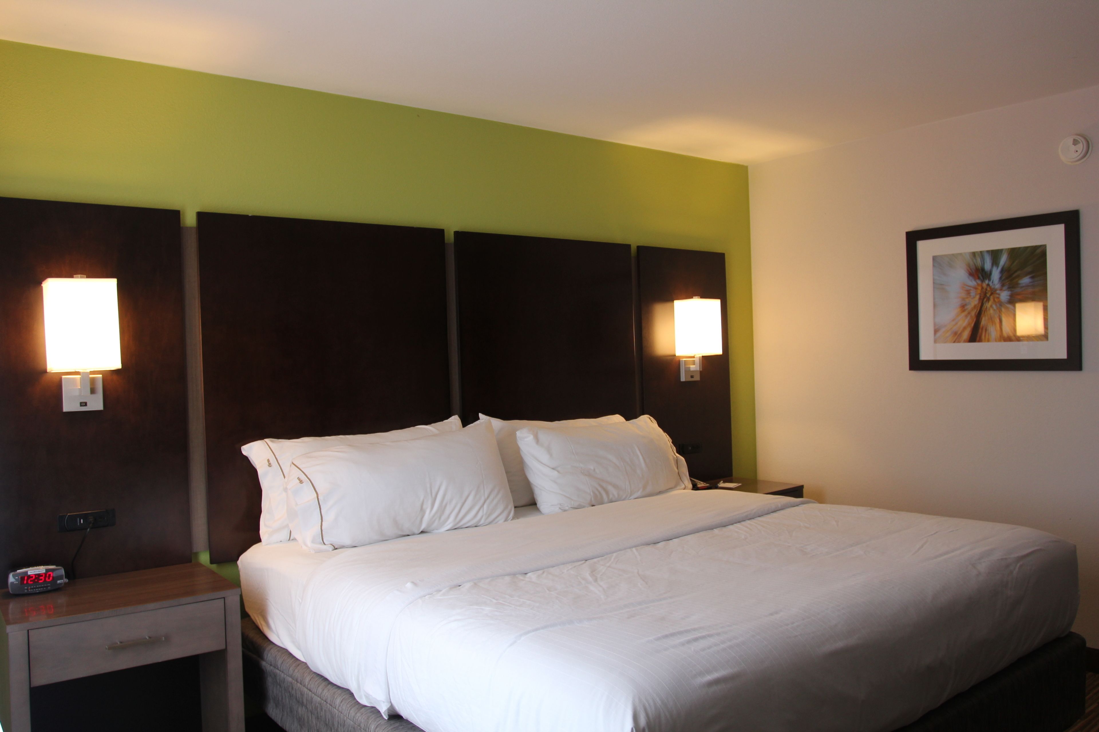 A hotel room with a large bed, two bedside tables with lamps, a nightstand with a clock, and a framed picture on the wall.