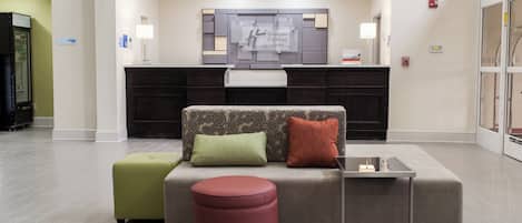 Un moderno vestíbulo de hotel con una recepción, un sofá con cojines, un pequeño reposapiés y una mesa de centro de vidrio.