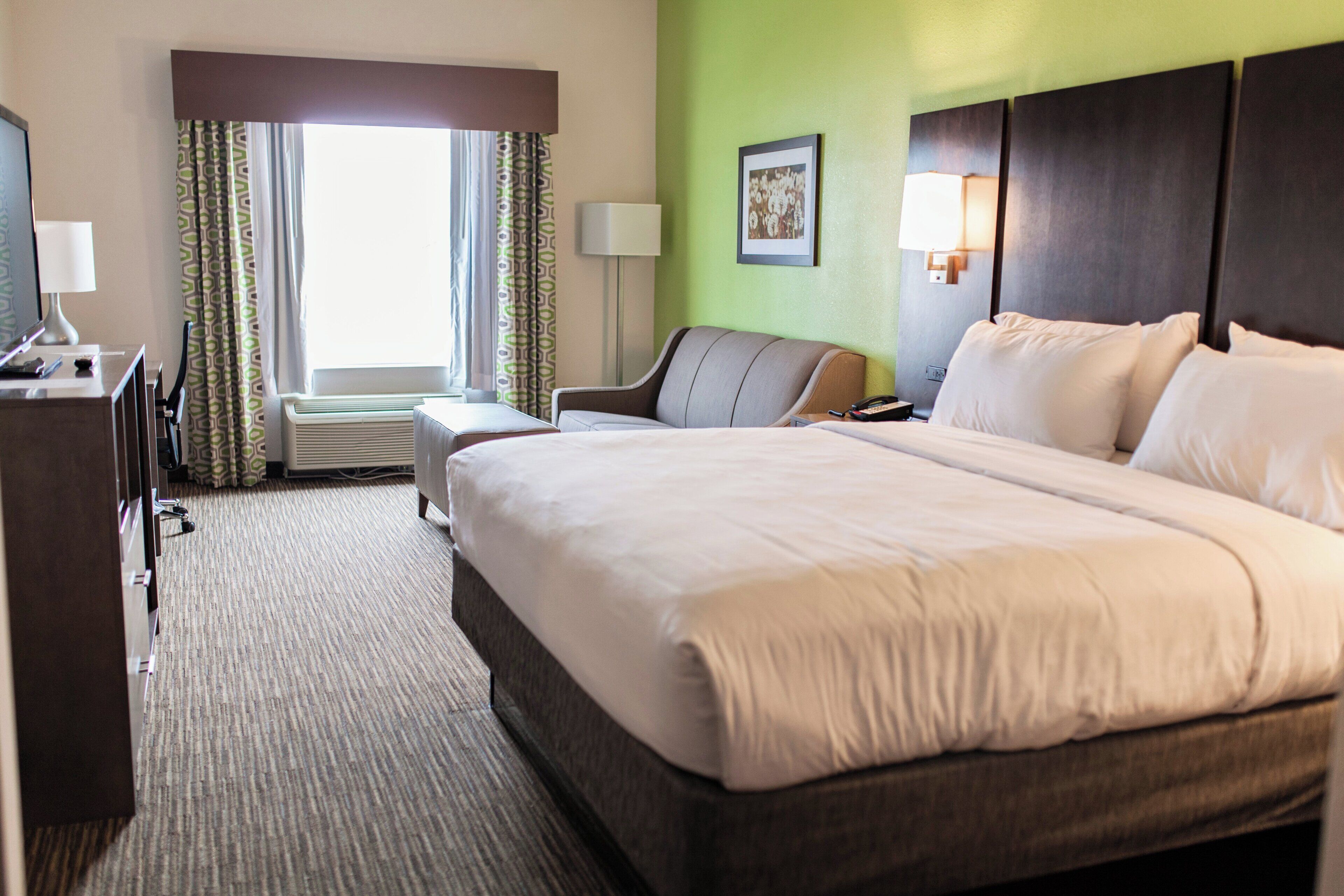 A hotel room with a large bed, a desk with a lamp, a chair, and a window with curtains.