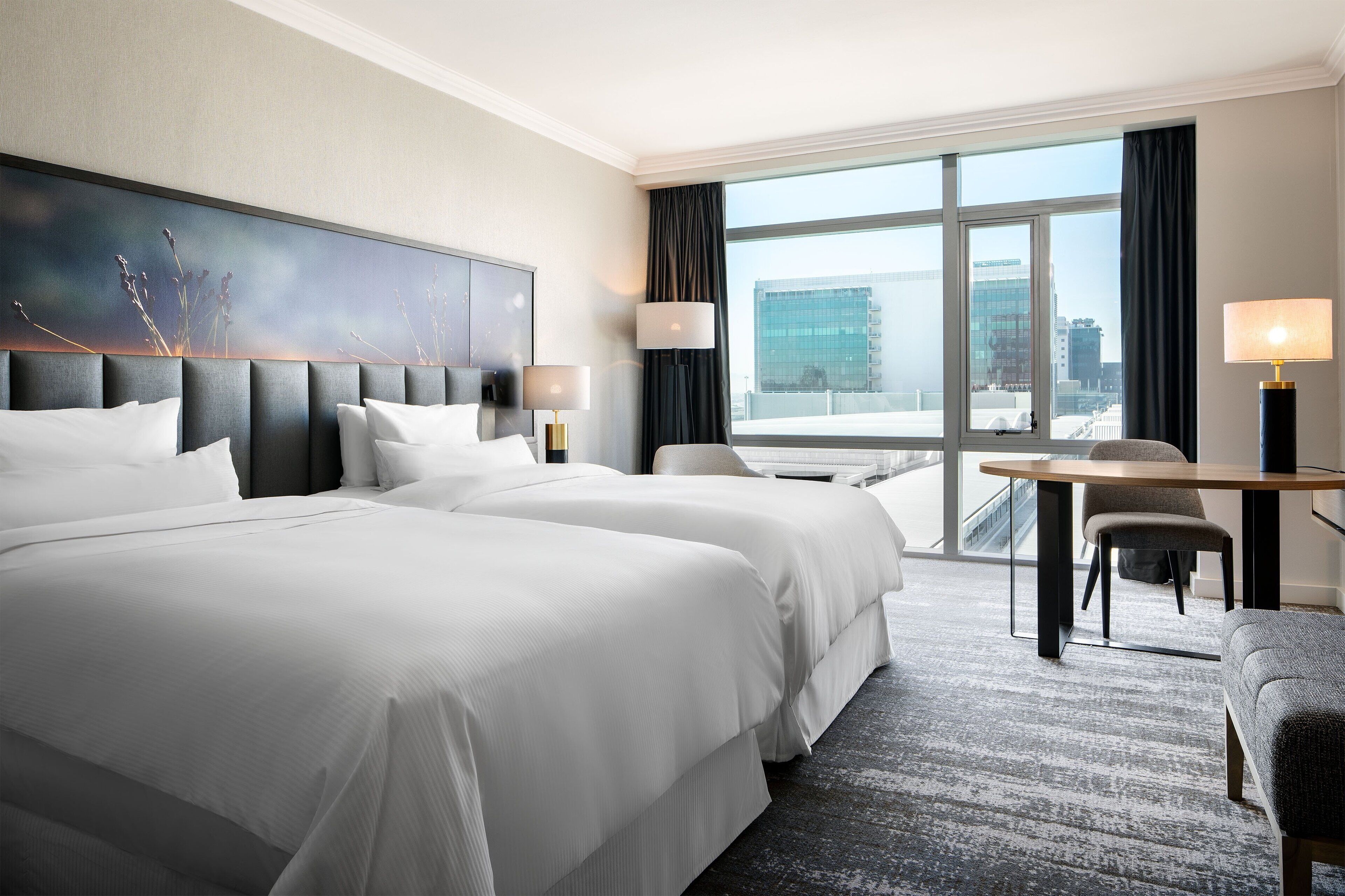 Habitación de hotel con dos camas grandes, un escritorio con silla y vistas a un paisaje urbano a través de amplias ventanas.