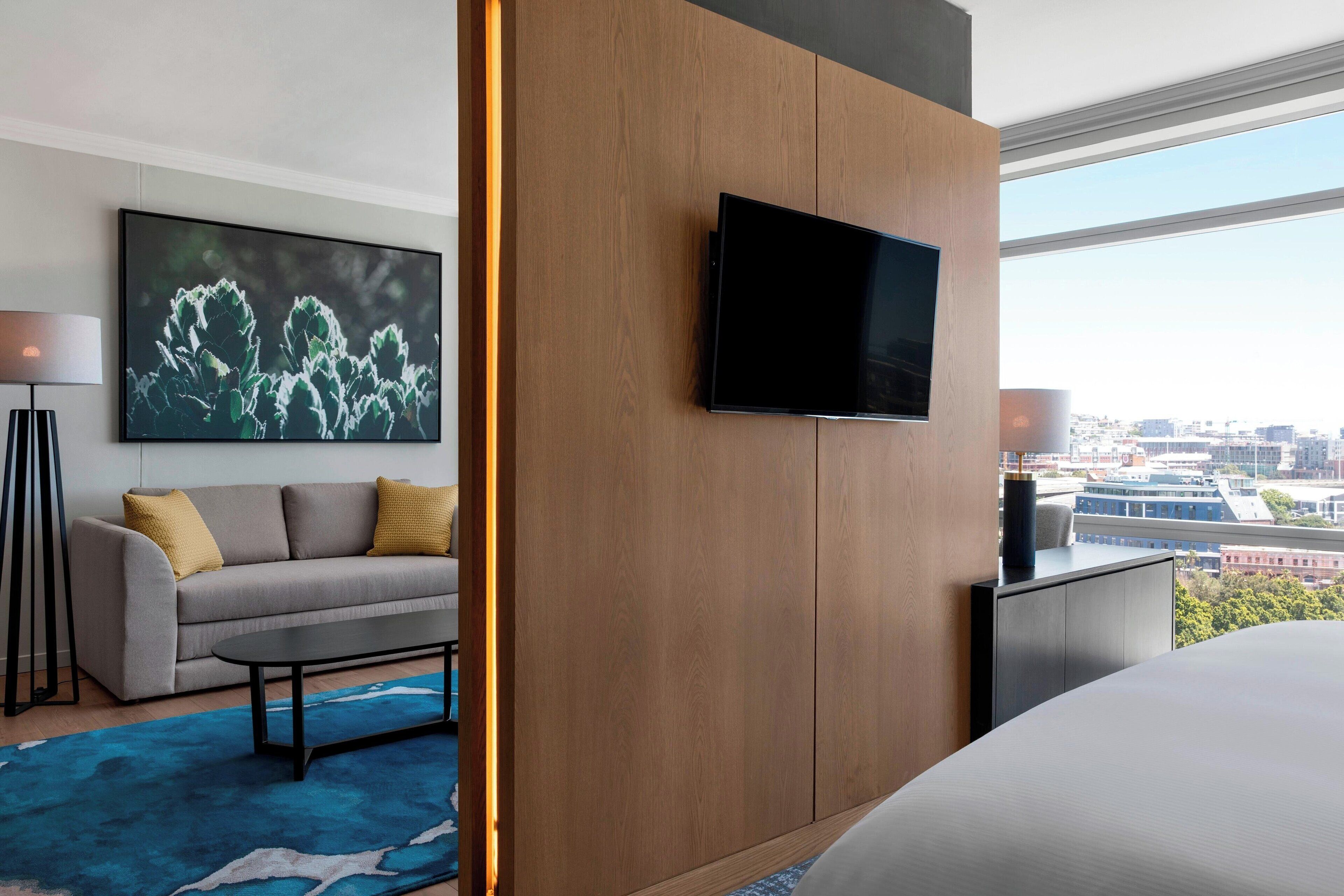 A modern hotel room with a large bed, a flat-screen TV mounted on a wooden panel, a sofa with cushions, a coffee table, and a city view through large windows.