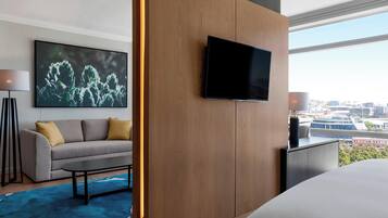 A modern hotel room with a large bed, a flat-screen TV mounted on a wooden panel, a sofa with cushions, a coffee table, and a city view through large windows.