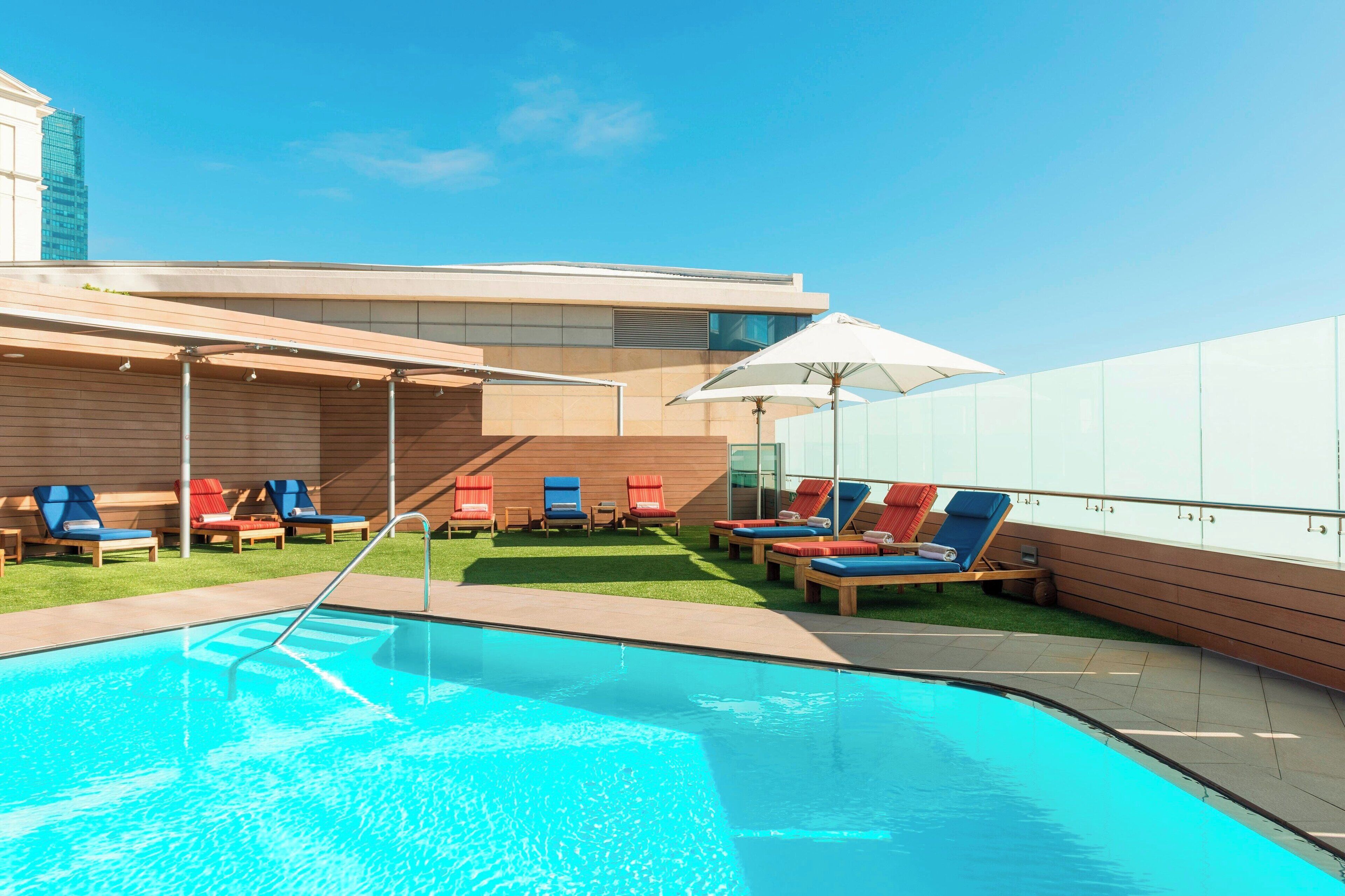 Un espace piscine moderne, doté de chaises longues et de parasols, entouré d’un bâtiment aux grandes fenêtres, avec un ciel dégagé.