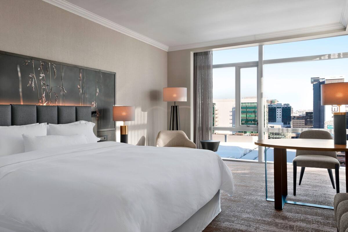 Een hotelkamer met een groot bed, een bureau met twee stoelen, uitzicht op de stad door grote ramen en een moderne lamp.