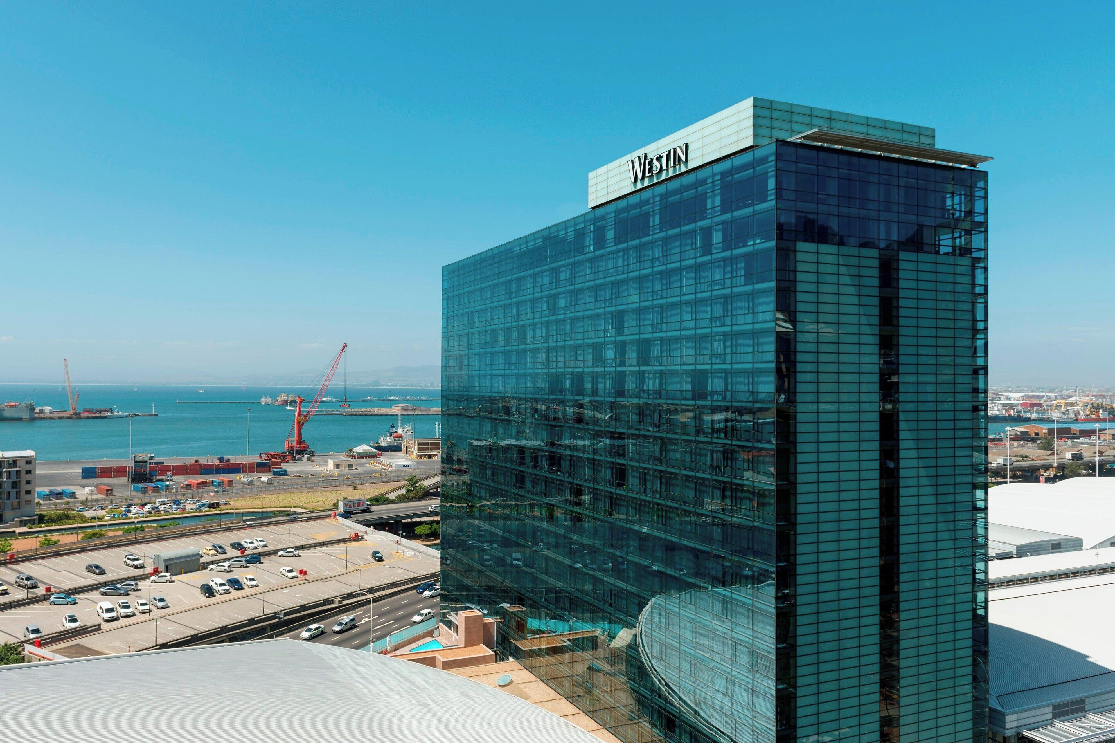 Un hotel Westin moderno con fachada de vidrio, situado cerca de un puerto con grúas visibles y un estacionamiento.