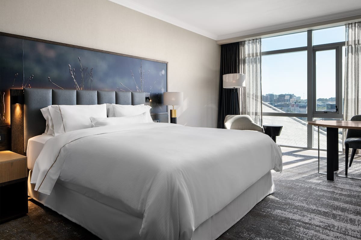 Een moderne hotelkamer met een groot bed, een bureau en uitzicht op de stad door grote ramen.