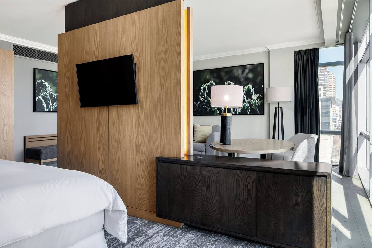 Een moderne hotelkamer met een groot bed, een houten scheidingswand, een flatscreen televisie, een ronde tafel met een lamp, en uitzicht op de stad door grote ramen.