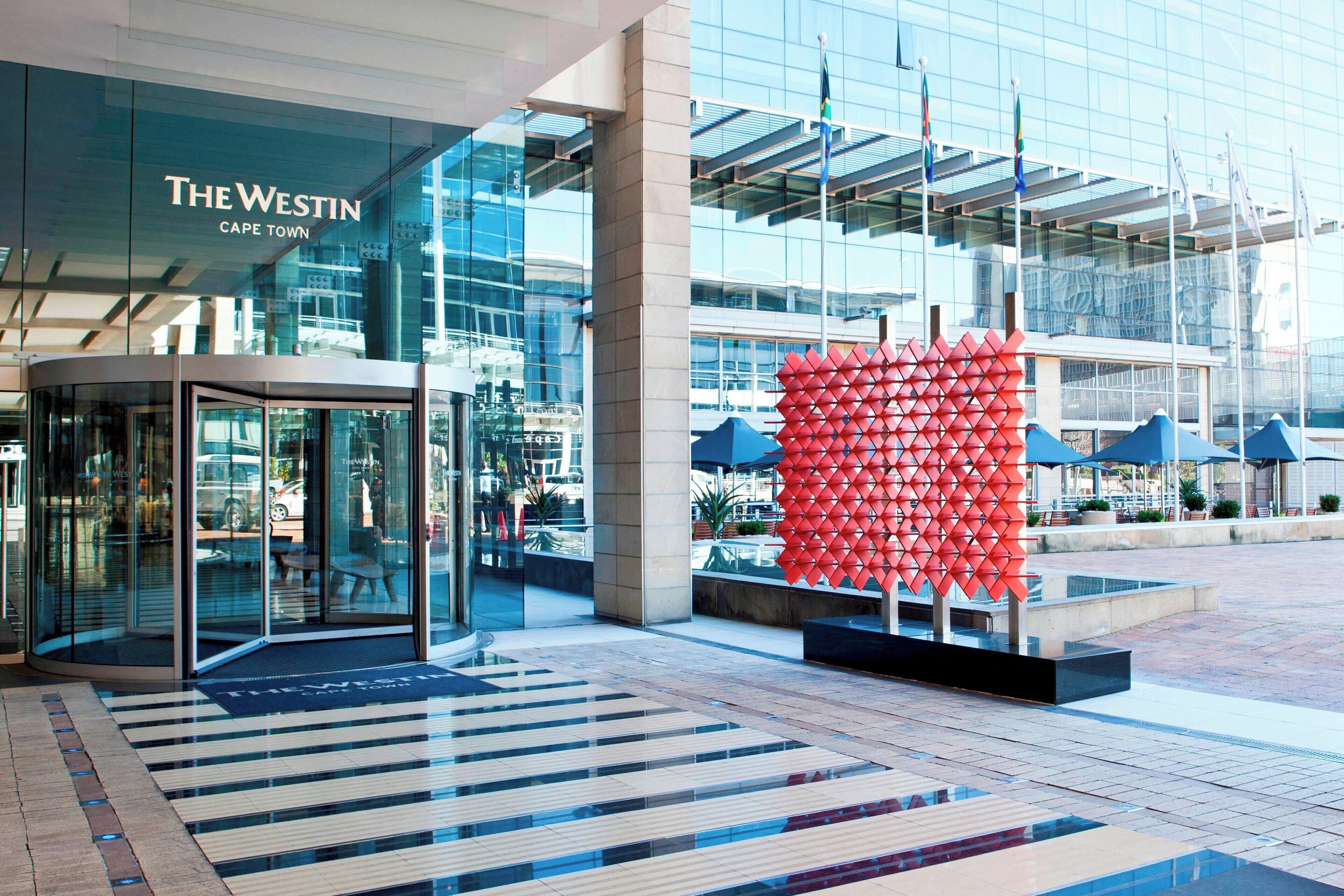 A modern hotel entrance with glass doors, a red geometric sculpture, and a tiled plaza.