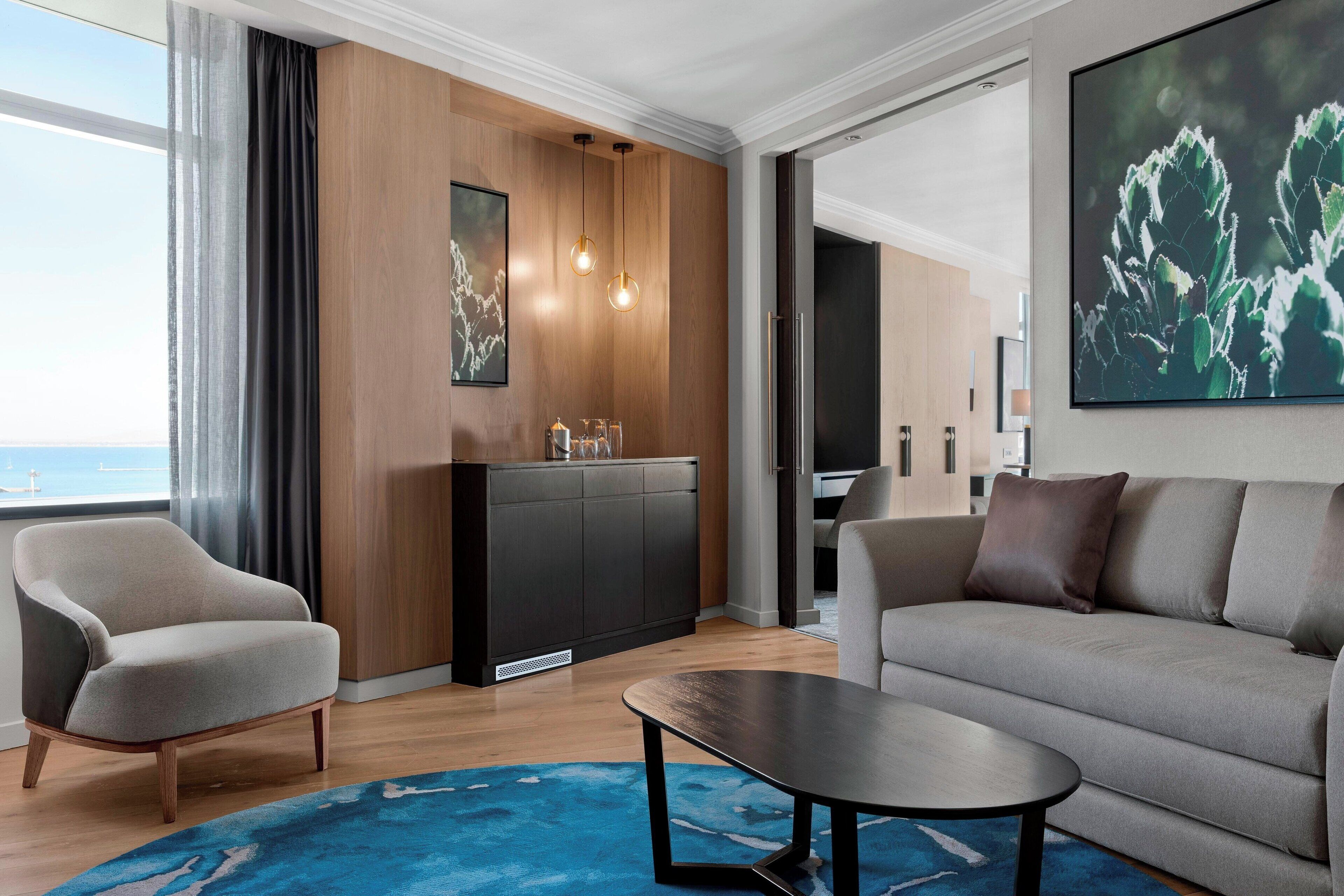 Uma sala de estar moderna com um sofá, poltrona e mesa de centro. Há um aparador de madeira com tampo de vidro e uma pintura na parede.