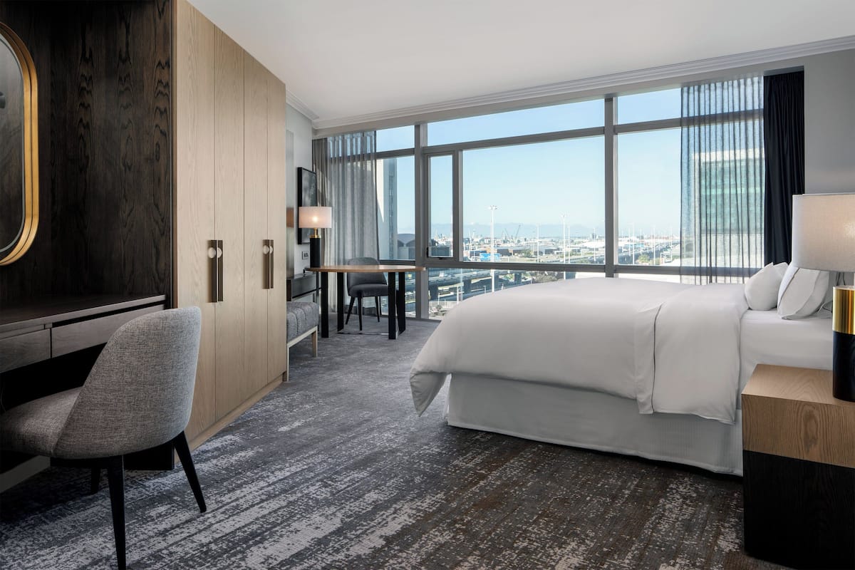 Hotelkamer met een groot bed, een bureau en een stoel. Er is uitzicht op de stad door grote ramen.