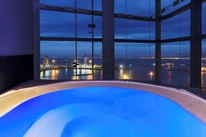 A modern indoor swimming pool with a panoramic view of a cityscape at dusk.