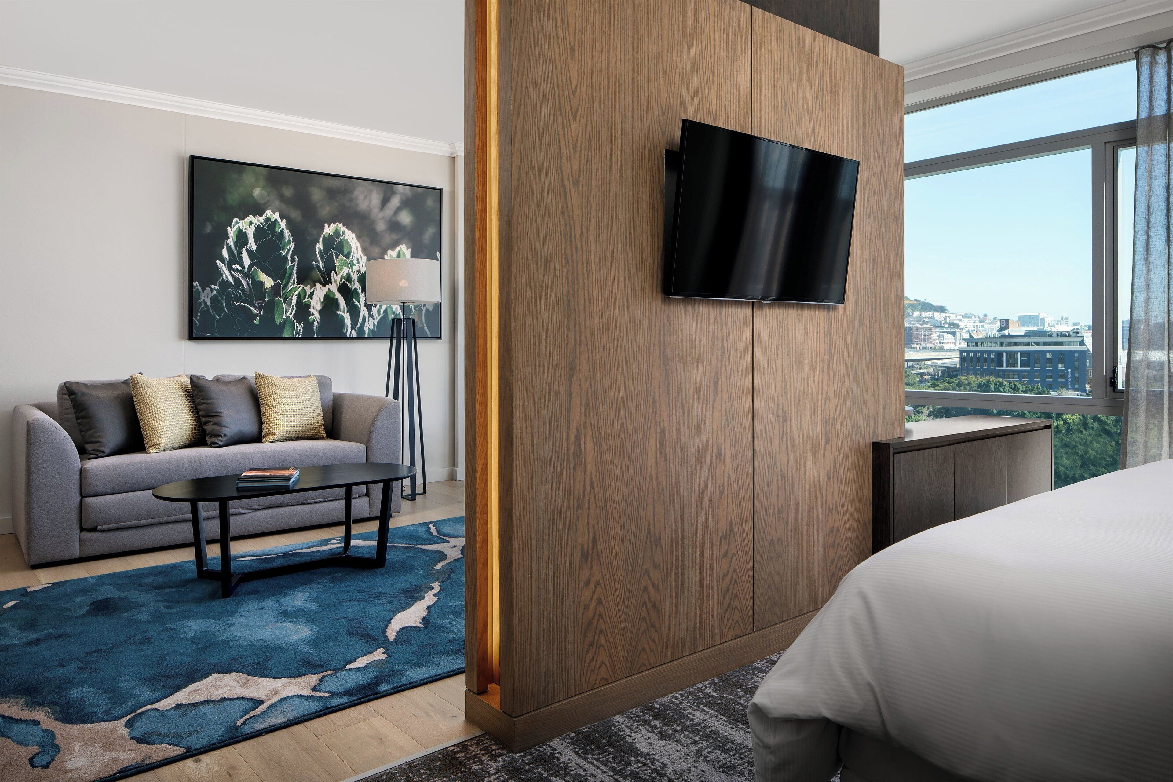 Quarto de hotel moderno com uma cama grande, um sofá, uma mesa de centro, uma TV fixada em um painel de madeira e vista para a cidade através de janelas amplas.