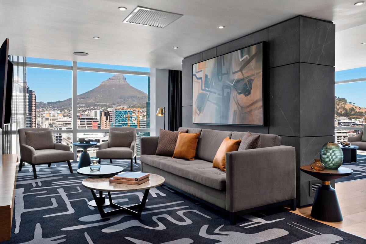 Een moderne woonkamer met een bank, fauteuils en een salontafel. Aan de muur hangt een groot abstract schilderij, en op de achtergrond is een stadsgezicht en een berg te zien.