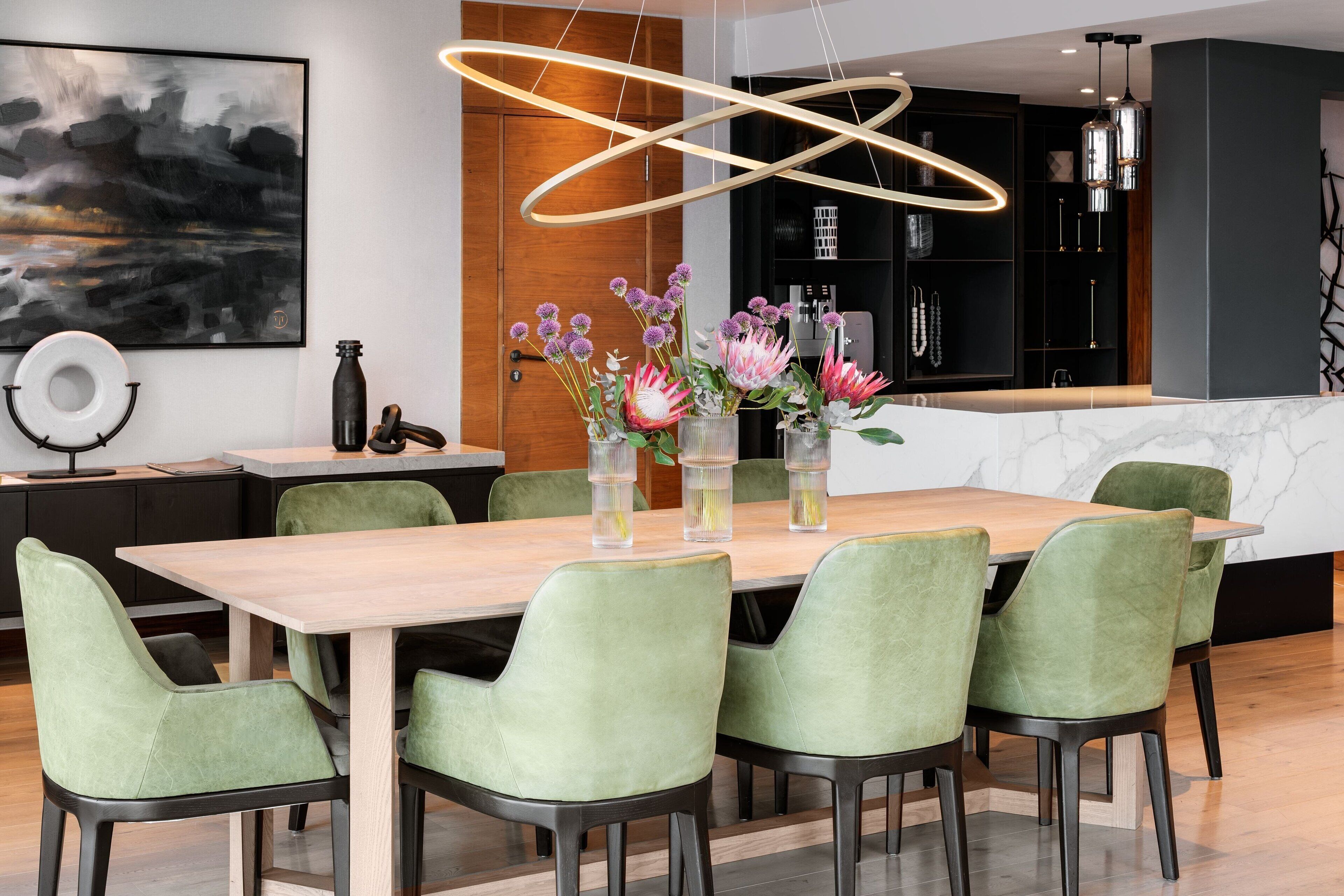 Uma sala de jantar moderna com mesa de madeira e cadeiras estofadas em verde, um armário preto com eletrodomésticos e uma grande pintura abstrata na parede.