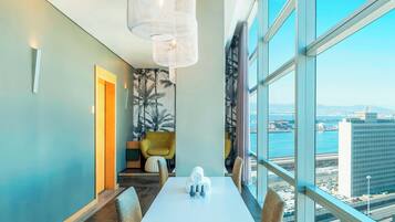 A modern hotel room with a large window offering a city view, a dining area with a white table, and a cozy seating area with a yellow chair.