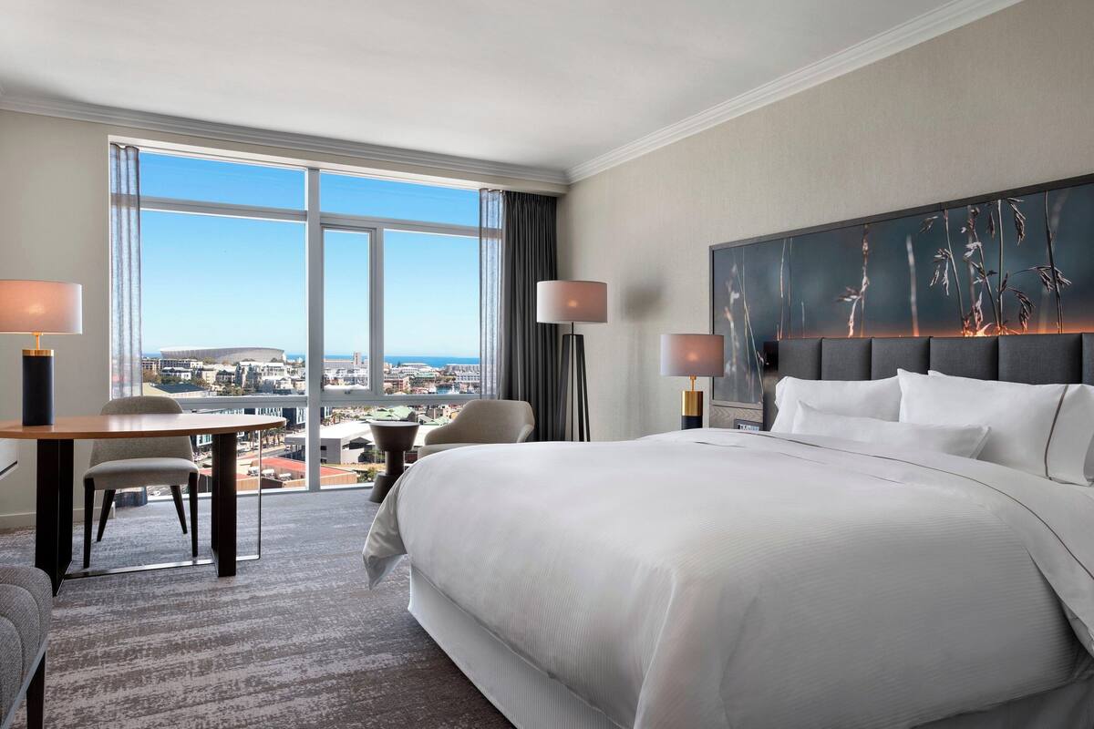 Een hotelkamer met een groot bed, een bureau en een stoel. Grote ramen bieden uitzicht op de stad.