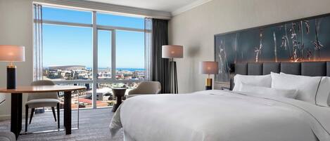 A hotel room with a large bed, a desk, and a chair. There is a view of the city through large windows.