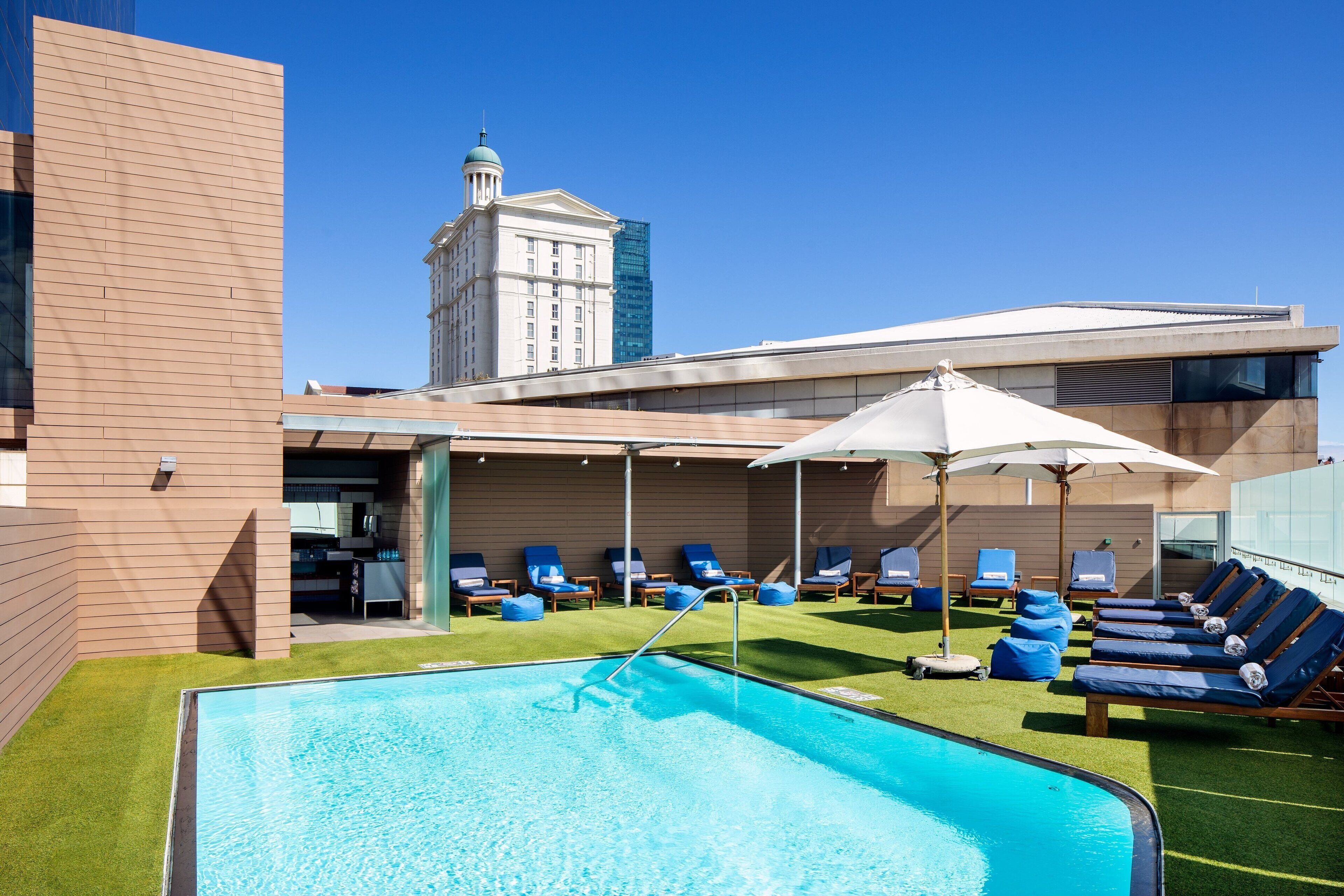 A rooftop pool area with lounge chairs and umbrellas, surrounded by modern buildings.