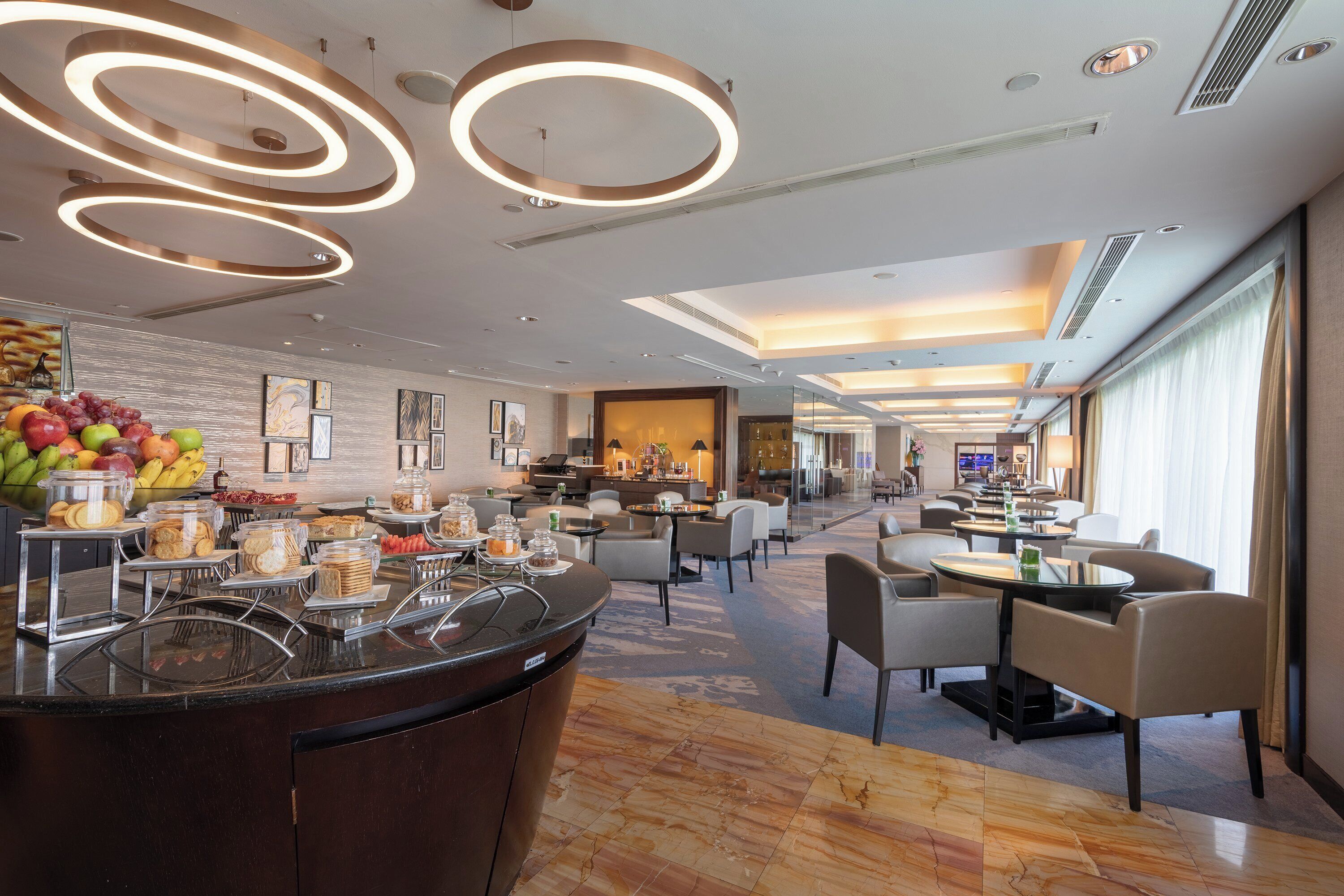 A modern hotel lobby with a central reception desk, a fruit display, and a seating area with tables and chairs.
