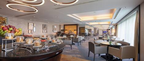 Un hall d’hôtel moderne avec un comptoir d’accueil central, un présentoir de fruits et un espace détente avec des tables et des chaises.