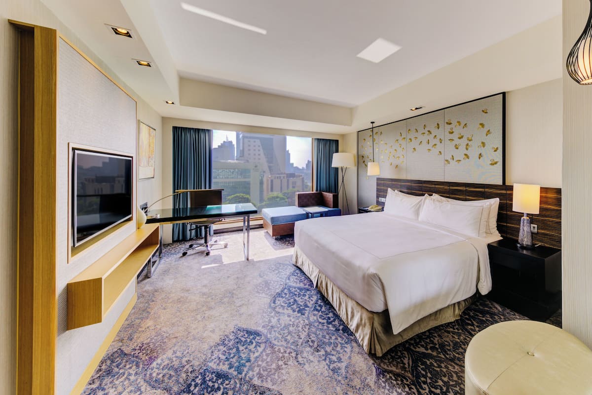Ett modernt hotellrum med en stor säng, en platt-TV, ett skrivbord med stol, en soffa och utsikt över stadsbilden genom fönstret.
