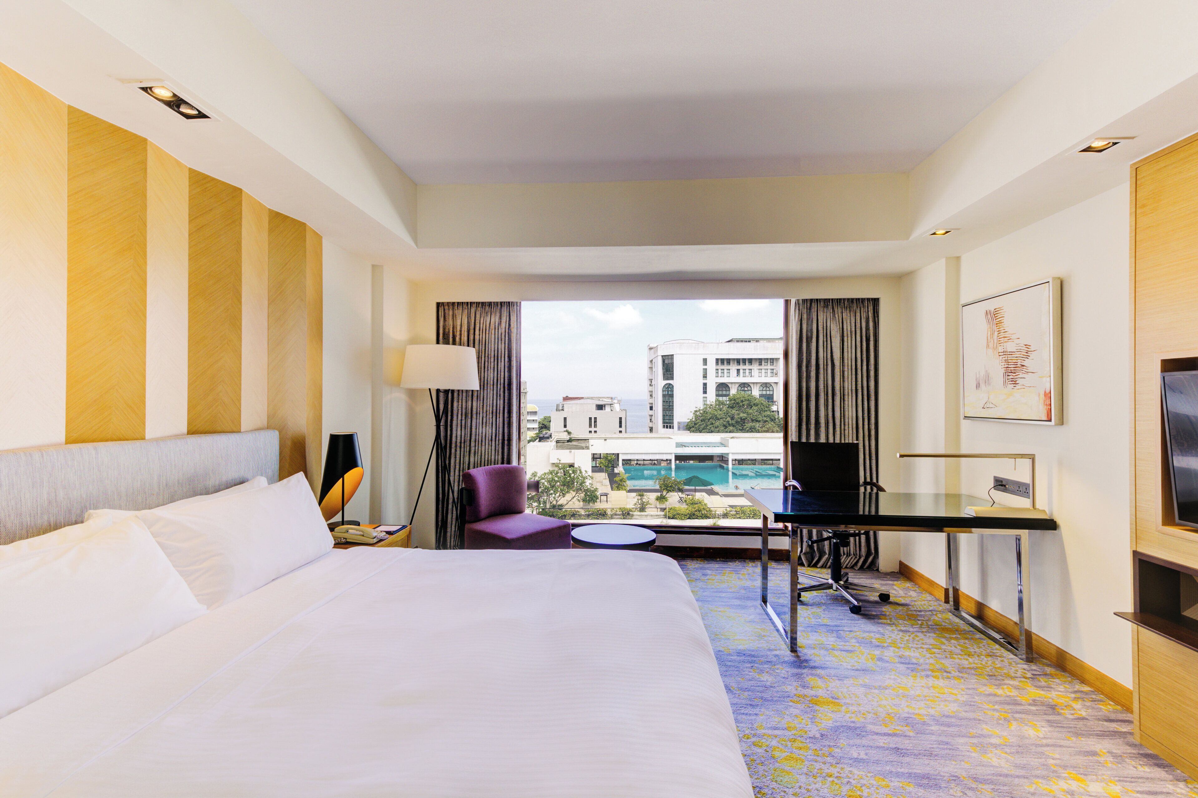 A hotel room with a large bed, a desk, a chair, a TV, and a view of buildings and a pool.
