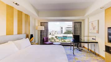 A hotel room with a large bed, a desk, a chair, a TV, and a view of buildings and a pool.