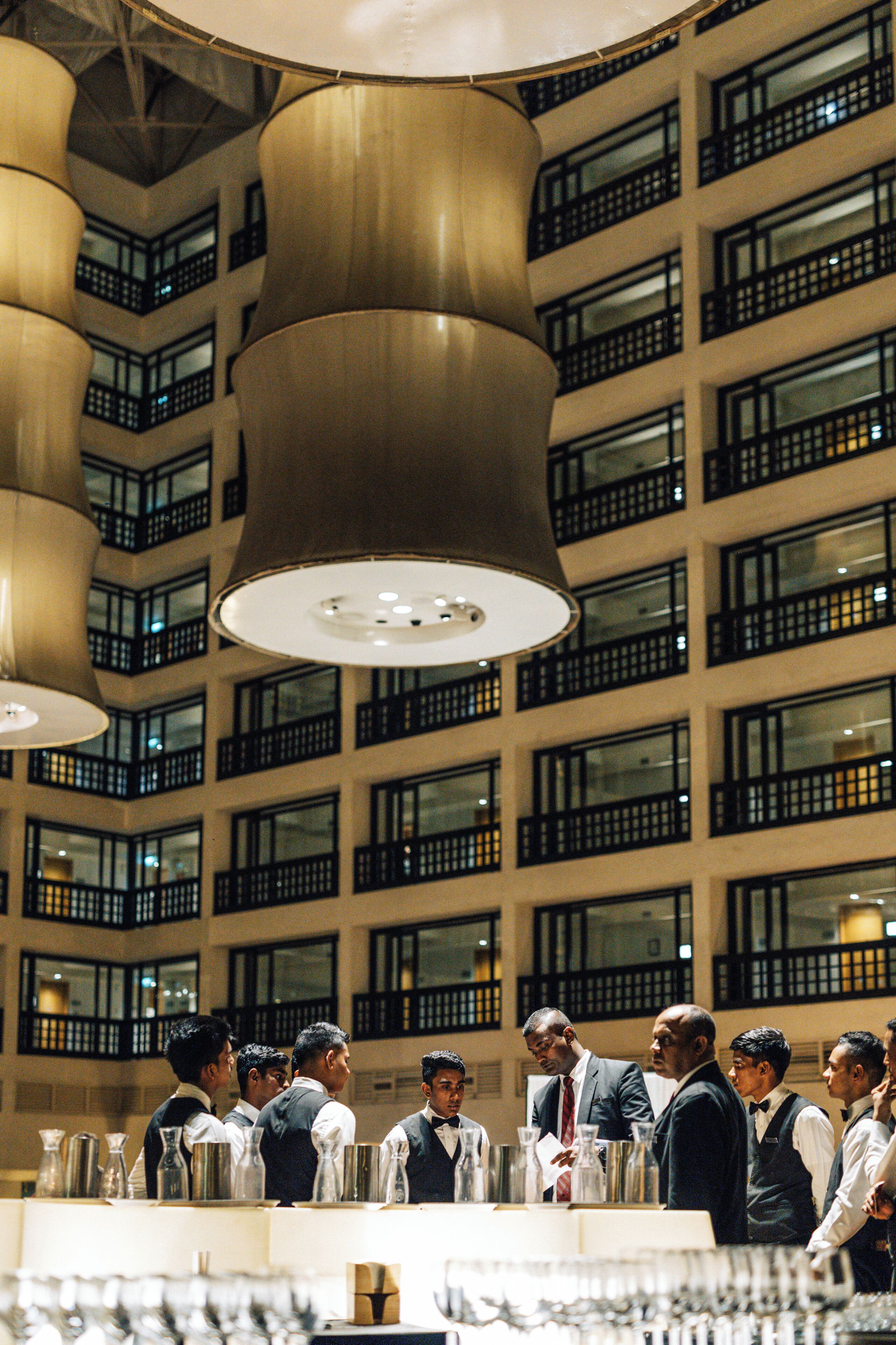 A group of waitstaff in a high-ceilinged dining area with multiple windows and hanging pendant lights.