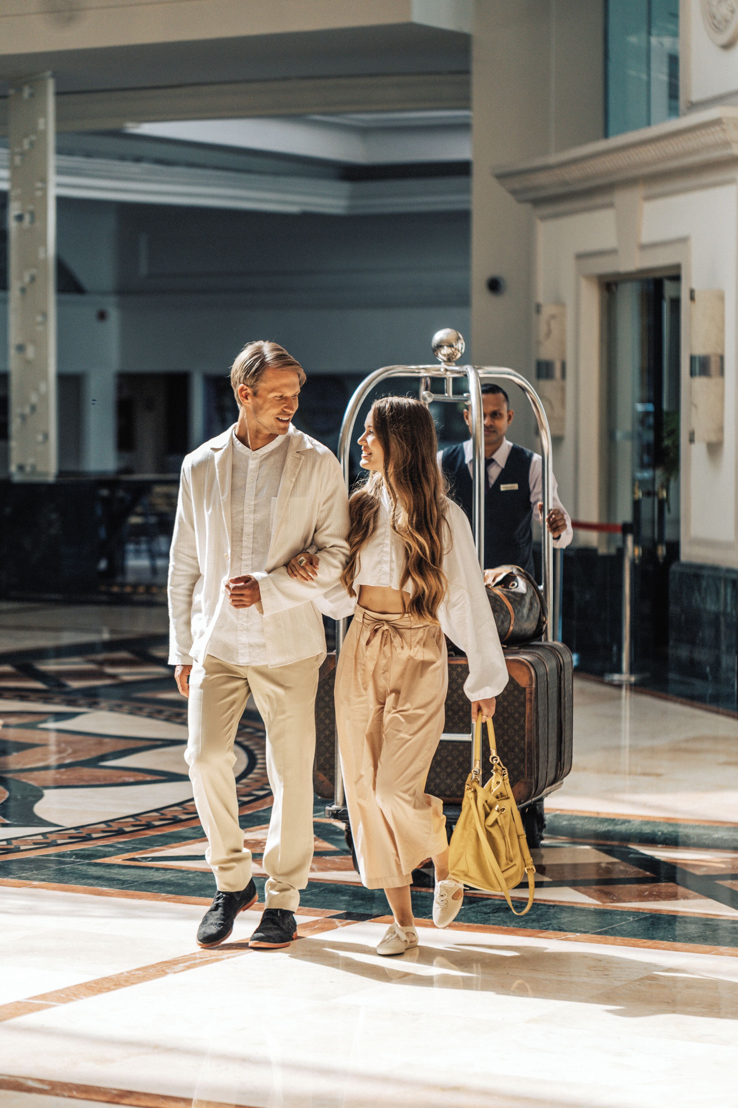 A couple walking towards a lift in a hotel lobby with a marble floor and a glass door in the background.