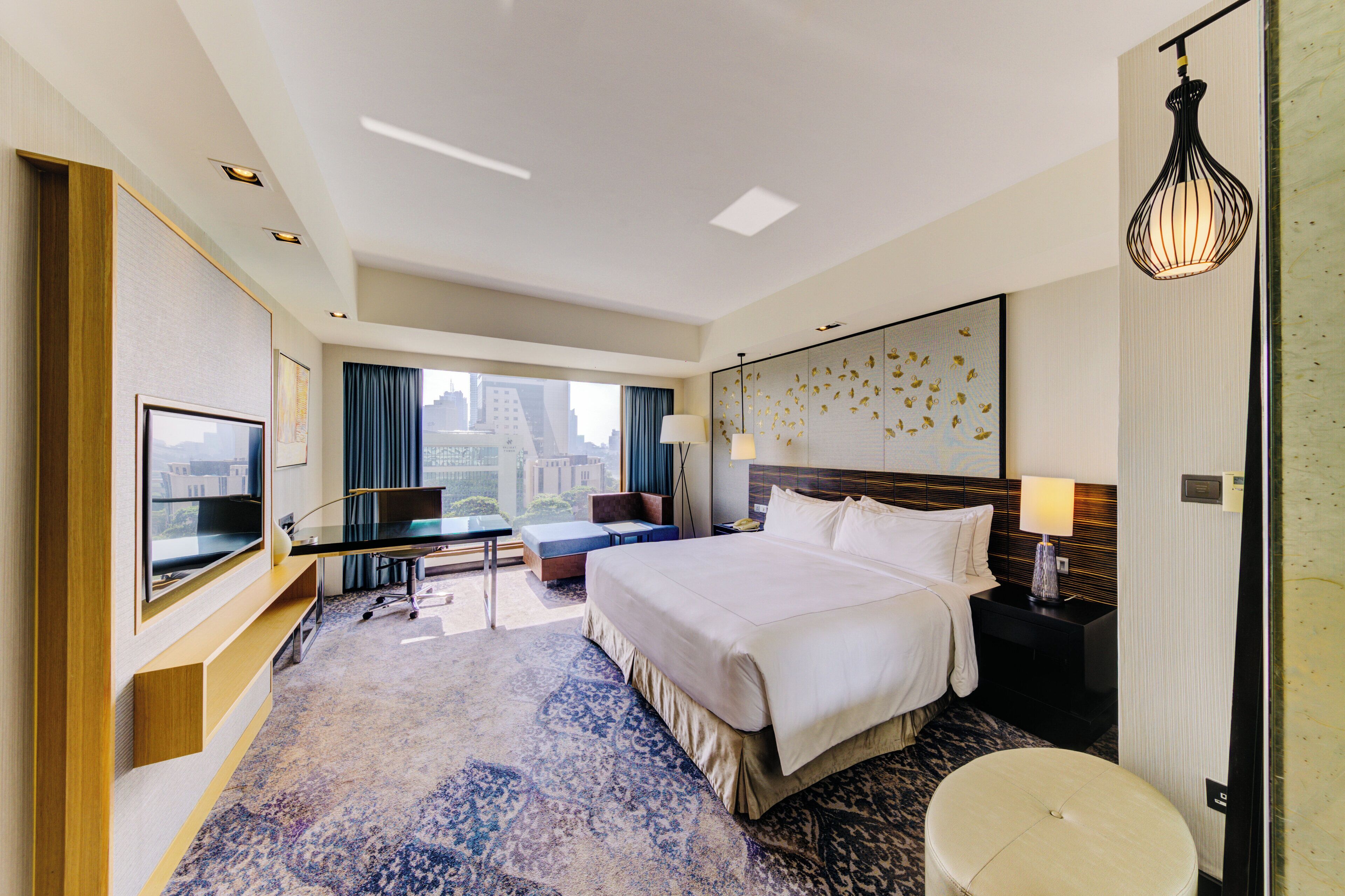 A modern hotel room with a large bed, a flat-screen TV, a desk, and a view of the city.