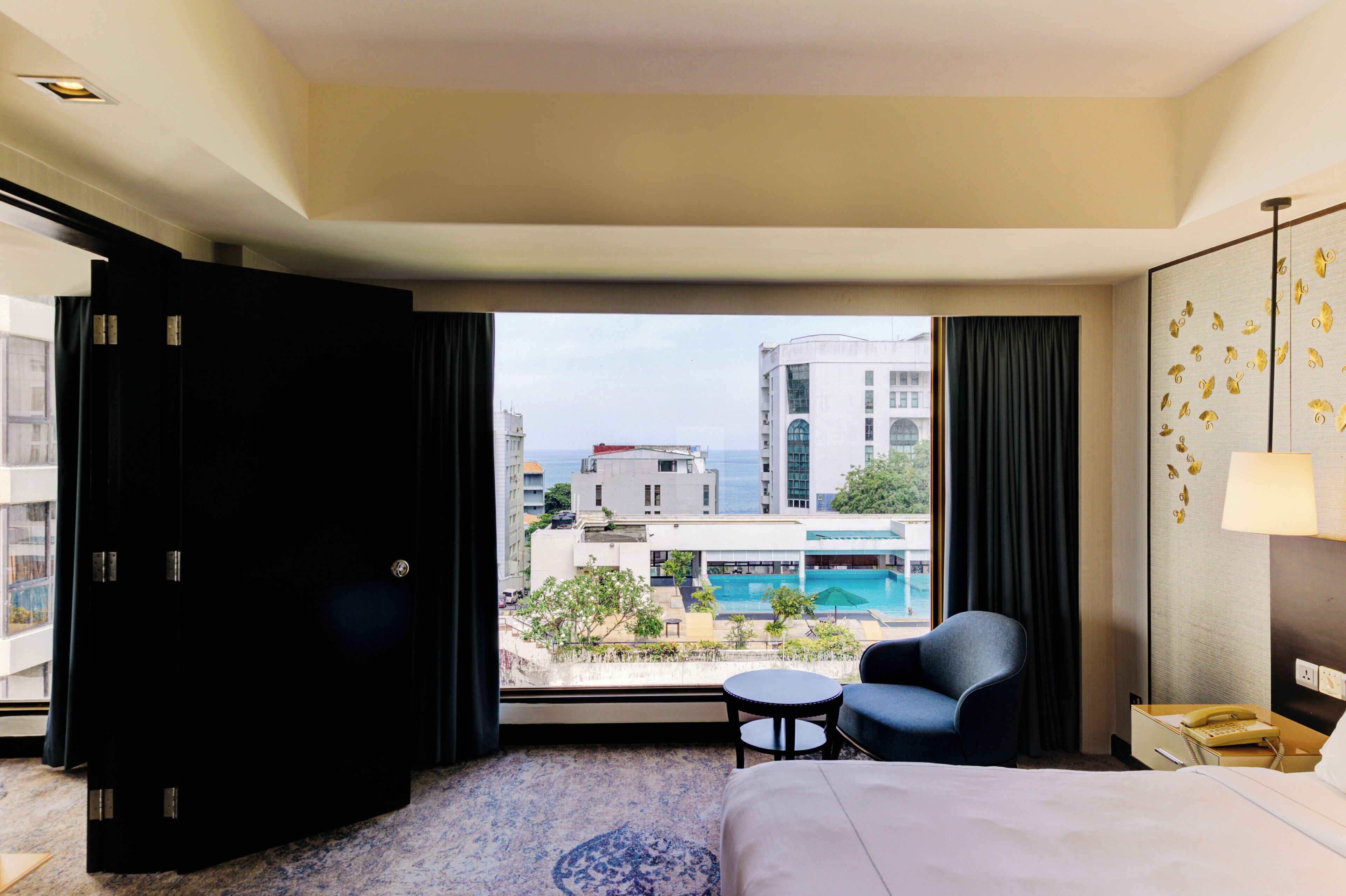 A hotel room with a large bed, a chair, a small table, and a view of a swimming pool and buildings.