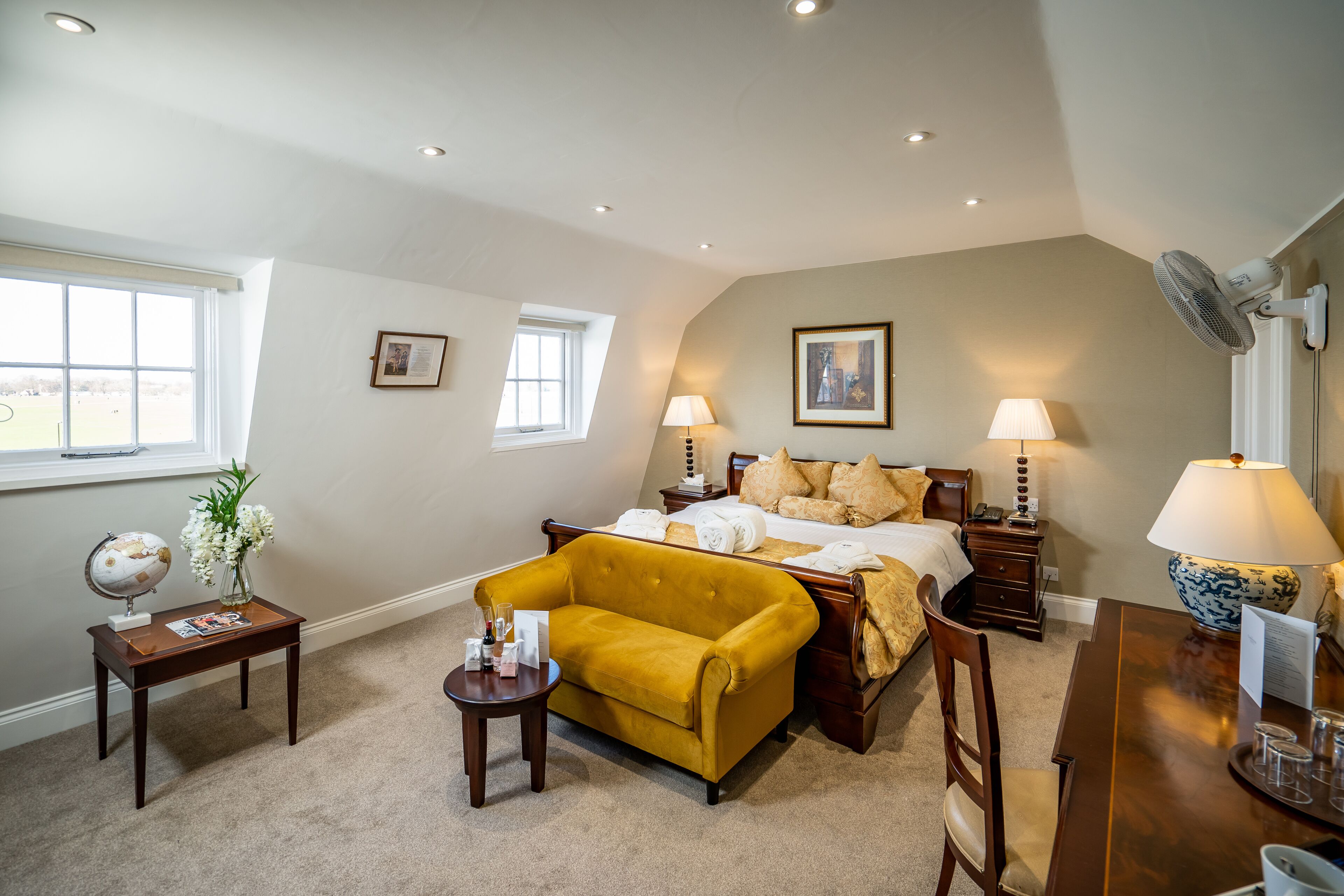 A hotel room with a bed, a yellow armchair, a wooden desk, a globe, and a vase with flowers.