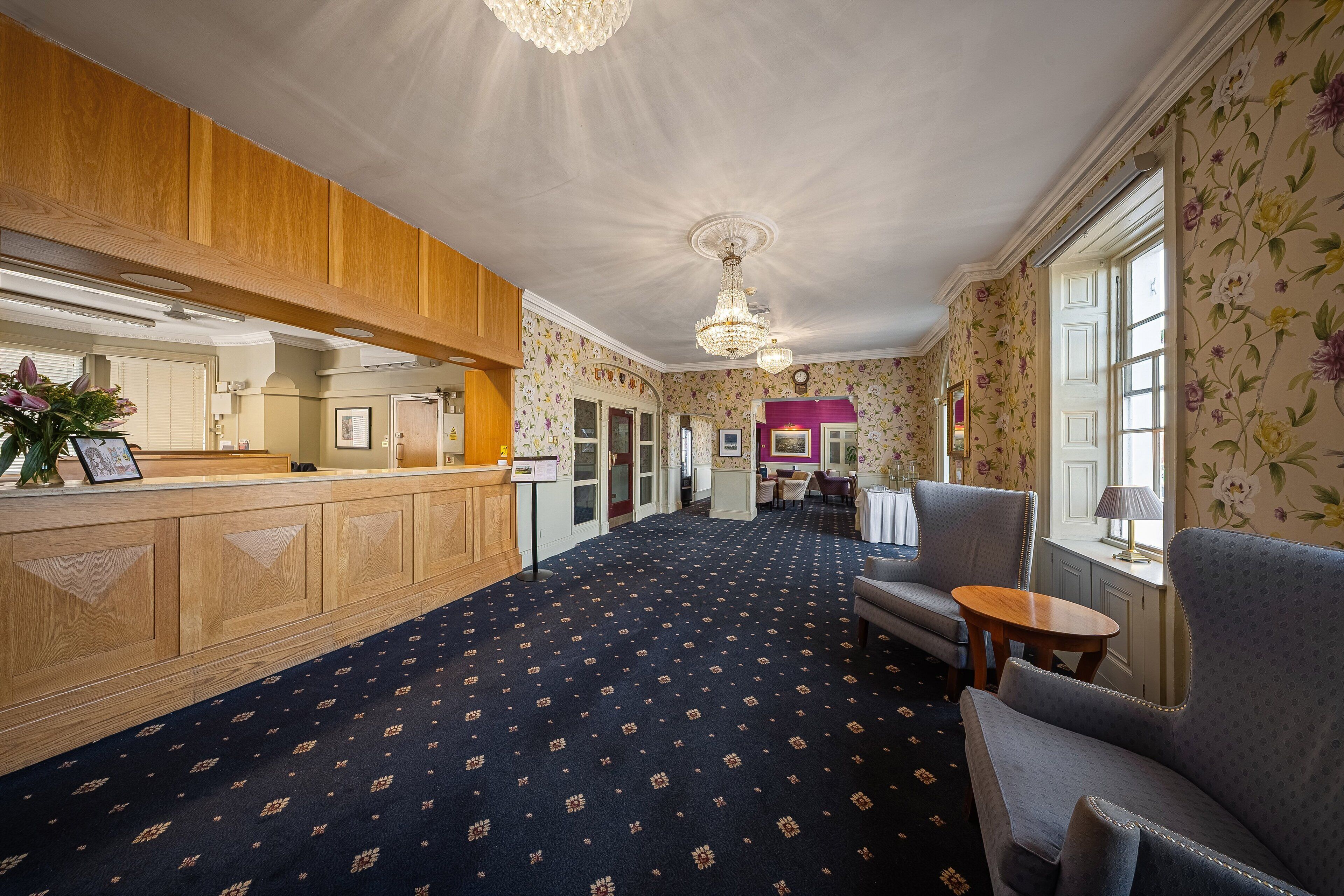 A hotel lobby with a reception desk, seating area, and floral wallpaper.