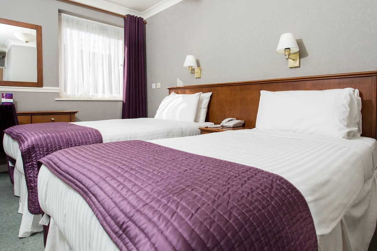 Een hotelkamer met twee bedden, een houten hoofdeinde, een nachtkastje met een telefoon en een raam met gordijnen.