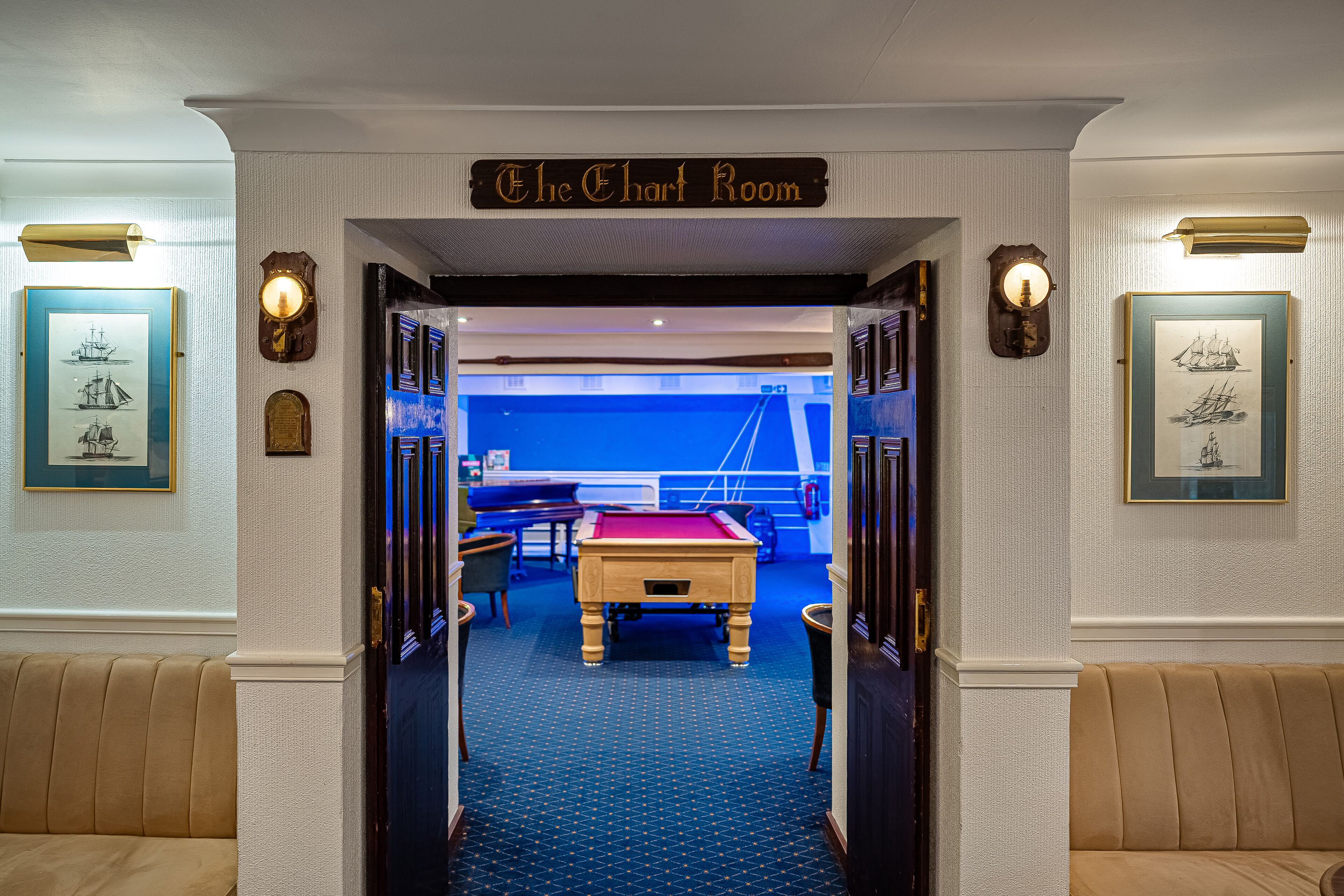 A billiards room with a pool table, framed nautical paintings, and a sign reading "The Chart Room".
