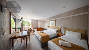 A hotel room with two beds, a desk, a chair, a desk lamp, a standing fan, and a window with a view of greenery. - The Clarendon Hotel - Blackheath (London)