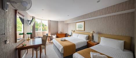 Habitación de hotel con dos camas, un escritorio, una silla, una lámpara de escritorio, un ventilador vertical y una ventana con vista a zonas verdes.