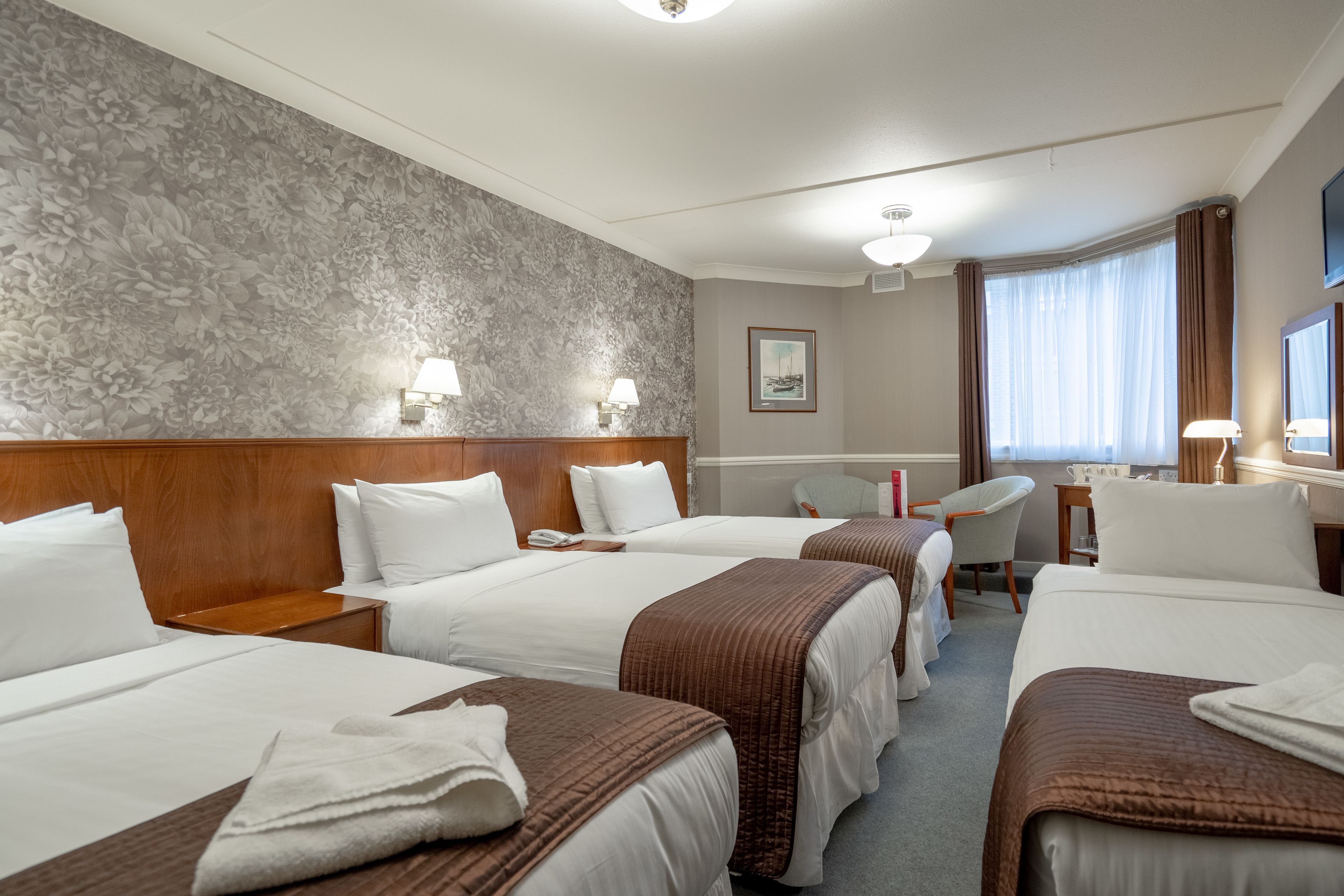 Habitación de hotel con tres camas, un escritorio, una silla y un ventanal con cortinas.