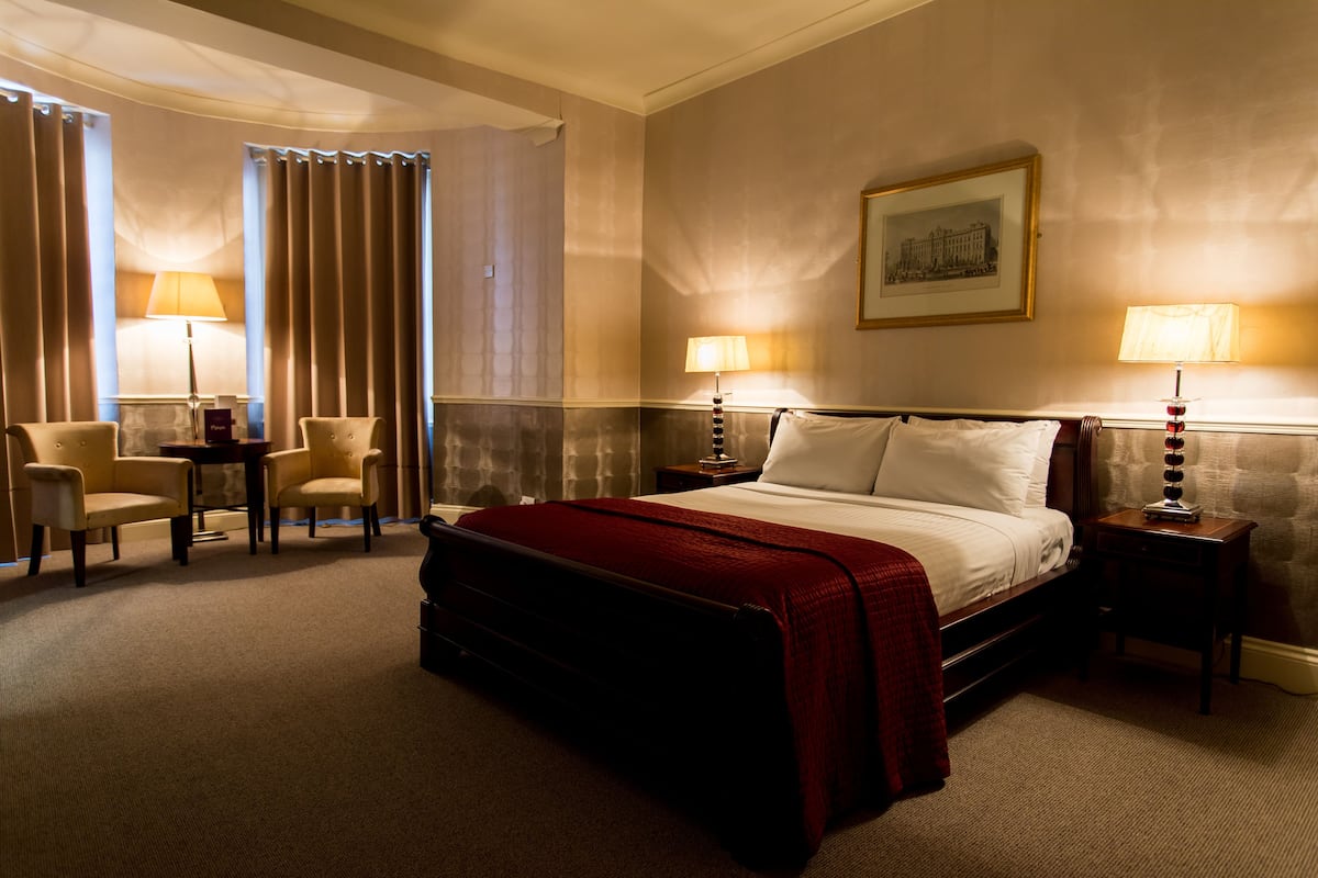Een hotelkamer met een groot bed, twee nachtkastjes met lampen, een stoel, een bijzettafeltje en een lijst met een afbeelding aan de muur.
