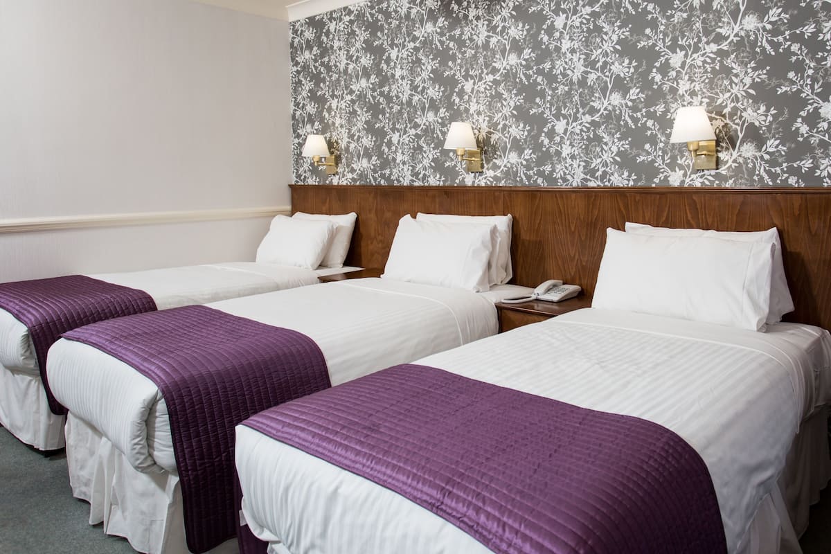 Een hotelkamer met drie bedden, elk voorzien van witte lakens en paarse dekbedovertrekken. Er zijn wandlampen op het hoofdeinde en een telefoon op het nachtkastje.