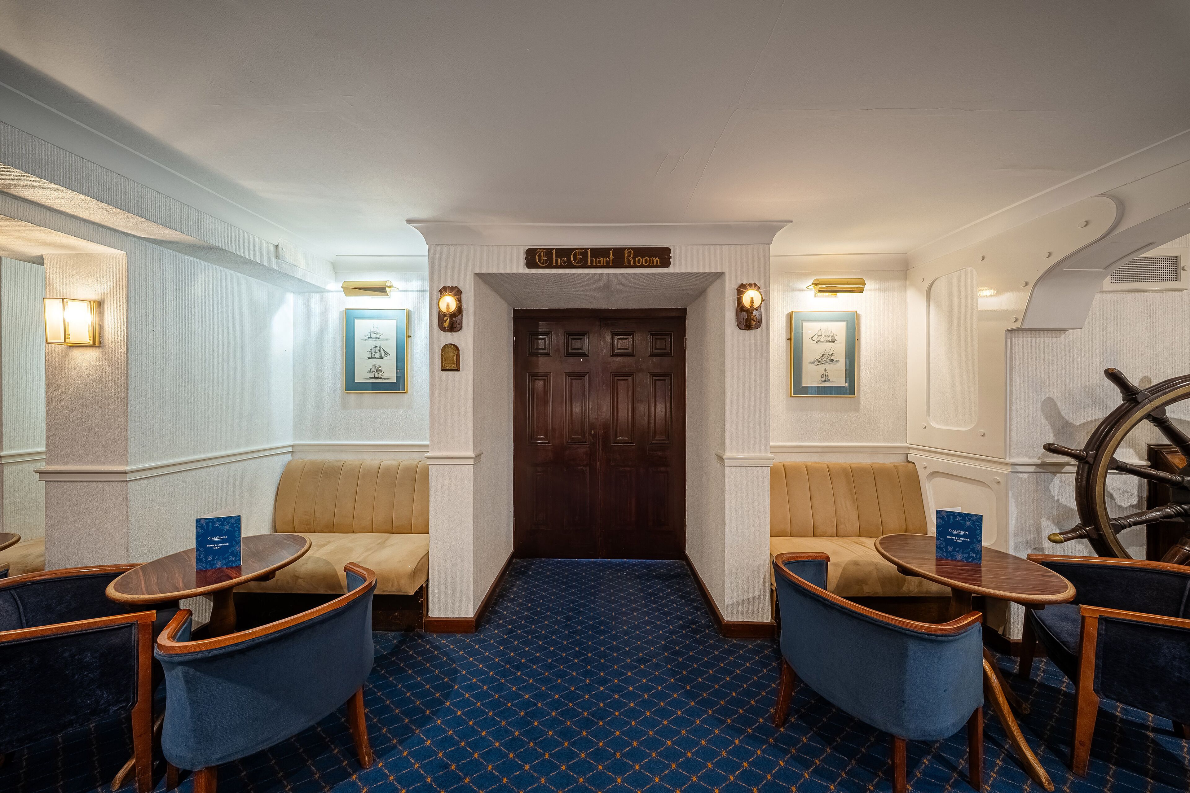 Una habitación con una puerta de madera con la leyenda "The Chart Room", alfombra azul estampada y dos sillones azules con estructura de madera.
