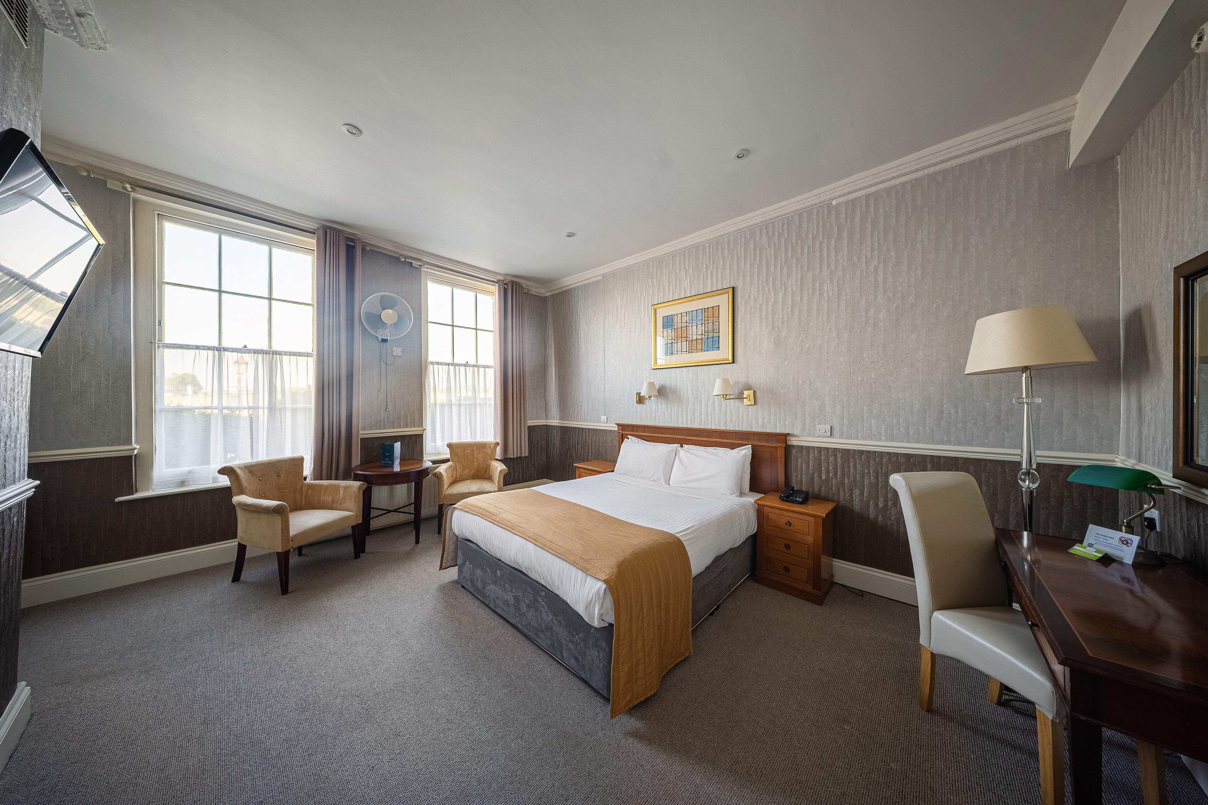A hotel room with a large bed, two armchairs, a desk, a standing lamp, a bedside table, a wall-mounted fan, and a window with curtains.